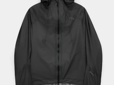 Front view of a black lightweight technical hooded jacket against a neutral background, featuring a full-length waterproof-style zipper and a structured hood. The fabric has a subtle grid texture with a slight sheen, complemented by articulated seam lines across the shoulders and sleeves. Adjustable hem drawcords and small tonal logos on the chest and sleeve emphasize a minimalist, performance-driven design