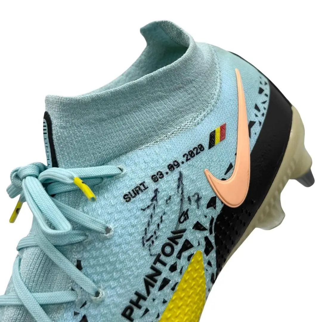 Close-up of a modern football boot in light blue knit with a sock-like collar, featuring a peach Swoosh, textured upper, graphic detailing, and visible “Phantom” branding with date and flag accents