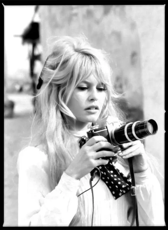 Black-and-white photograph of Brigitte Bardot holding a vintage camera, her tousled hair and soft gaze capturing a candid moment of 1960s cinematic elegance and off-duty intimacy
