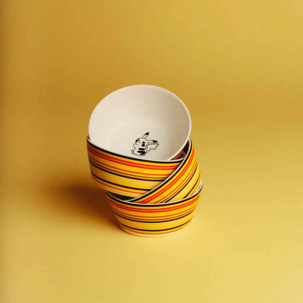 A stack of ceramic bowls arranged in a slightly tilted formation, each featuring bold horizontal stripes in yellow, orange, red, and black. The top bowl reveals a small black-and-white Pikachu illustration at its base. Set against a warm yellow background, the composition emphasizes the vibrant color bands and playful character detail with a clean, minimal aesthetic