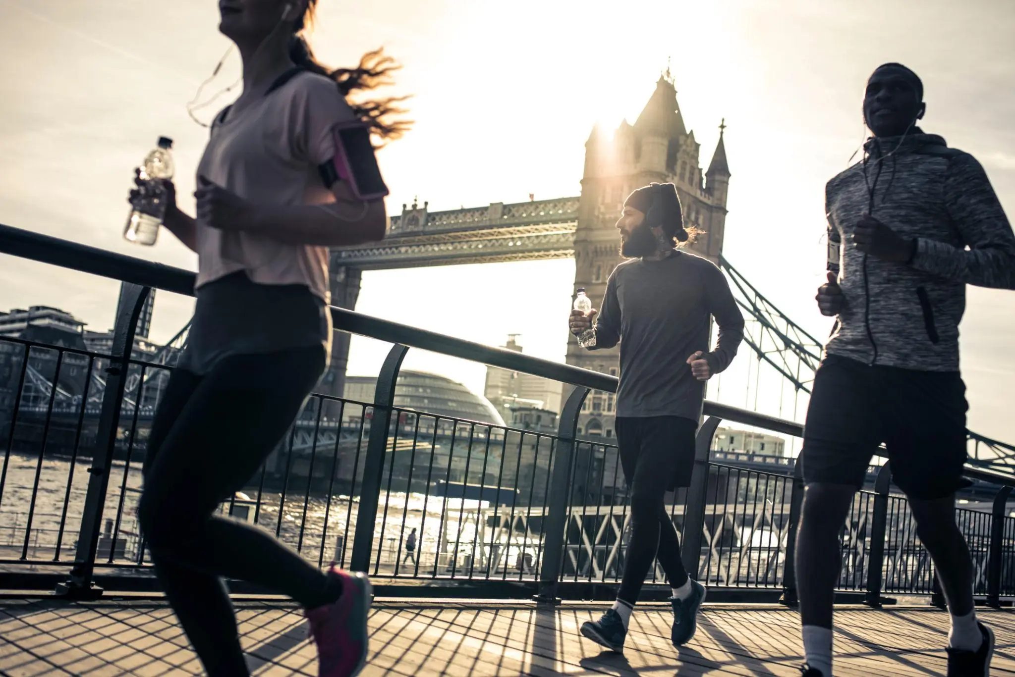 Three runners move along a riverside path near London’s Tower Bridge at sunrise, backlit by warm light, wearing athletic gear and headphones while holding water bottles, with the Thames and historic architecture in the background