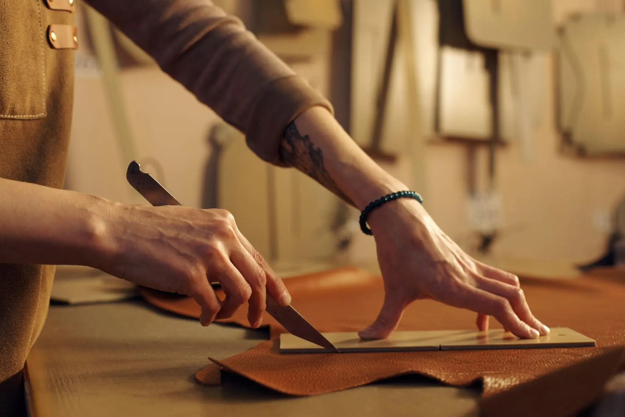 Close-up of hands cutting a piece of brown leather with a knife along a straightedge on a worktable, suggesting a craft or atelier setting