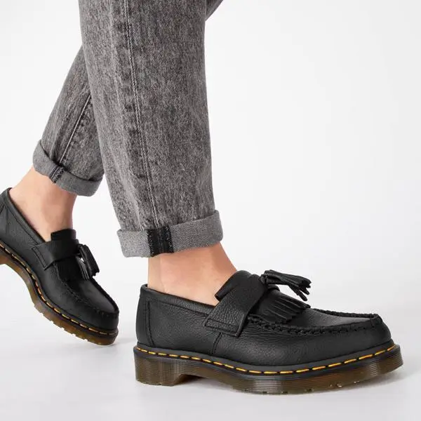A pair of black leather tassel loafers worn with cuffed grey jeans, featuring a textured grain finish, classic apron stitching, and a chunky translucent sole with signature yellow welt stitching
