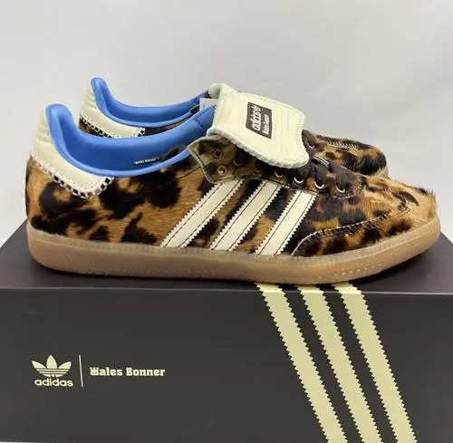 Low-profile shoe featuring a pony hair upper in a bold leopard print pattern, accented by three cream leather stripes along the side. The shoe includes a fold-over tongue with co-branded detailing, brown laces, and a translucent gum sole. Interior lining appears in a contrasting light blue. The pair is displayed on a black shoebox with co-branded adidas and Wales Bonner logos and signature triple stripe graphic