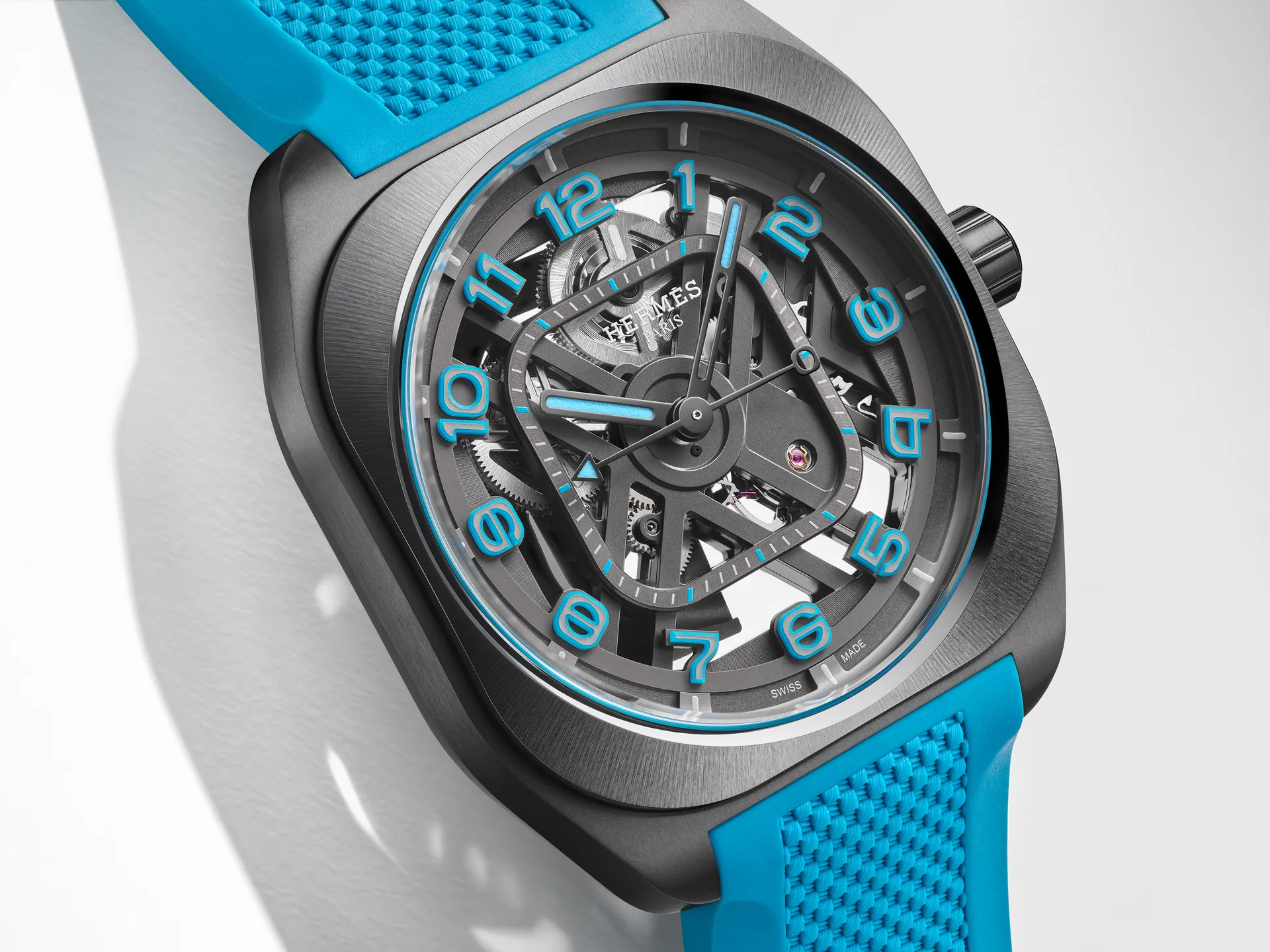 Angled close-up of a Hermès H08 Squelette watch in brushed titanium with an openworked dial exposing the mechanical movement, featuring bold turquoise Arabic numerals, matching luminous hands, and a textured turquoise rubber strap against a clean white background