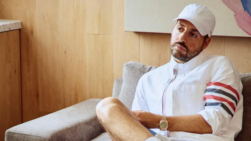 Ronnie Fieg, founder of Kith, sitting relaxed in a modern interior wearing a white Thom Browne shirt, white cap, and gold watch