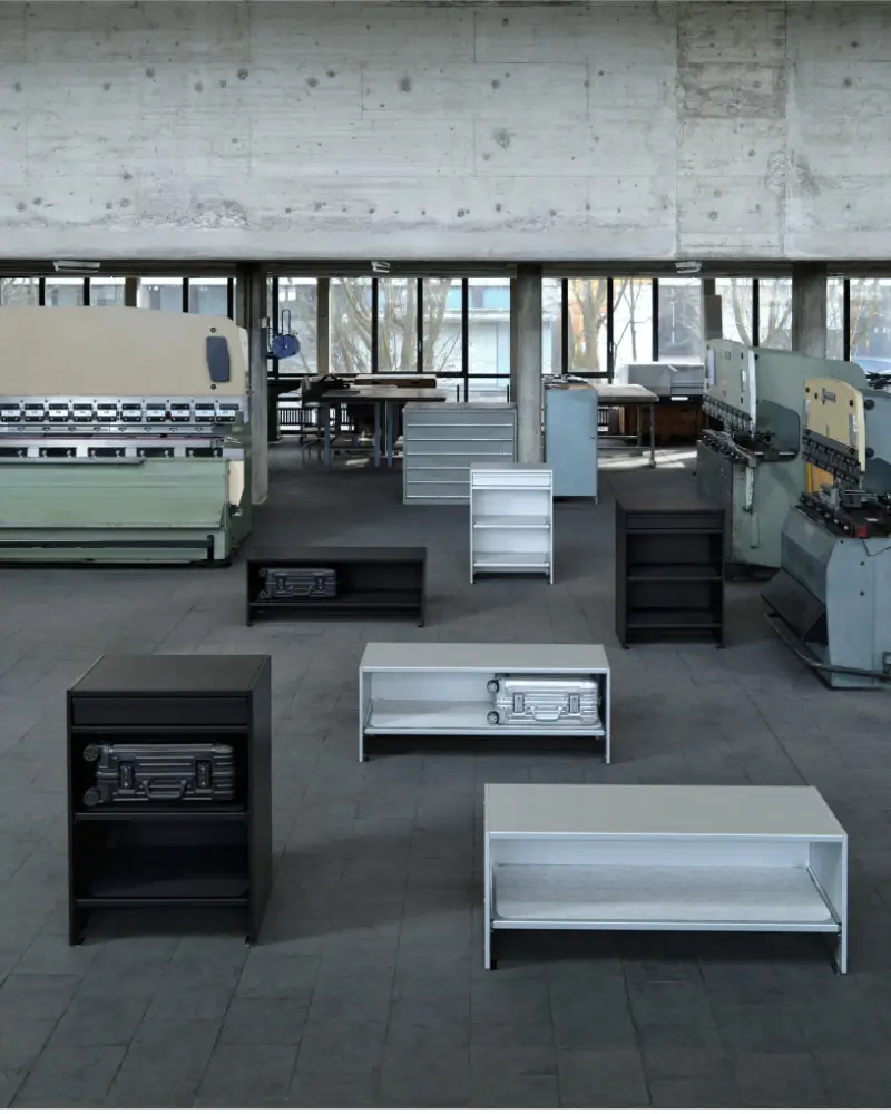 Rimowa x Lehni furniture installation set within an industrial workshop, featuring minimalist aluminum benches and storage units in black and silver, each designed to house suitcases, emphasizing precision engineering, modular design, and the translation of travel objects into architectural furniture forms