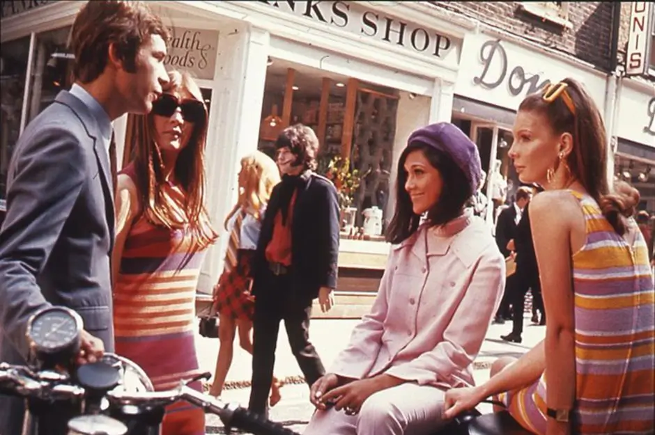 1960s London street scene with young people in colorful mod fashion gathered outside shops, reflecting the vibrant youth culture and style of the Sgt. Pepper era