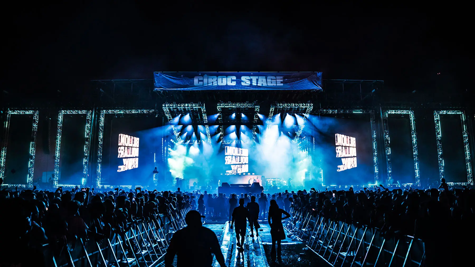Wide-angle view of a massive festival stage labeled “CÎROC STAGE,” illuminated with blue lighting and large LED screens, with a dense crowd gathered behind barricades and a central walkway leading toward the stage