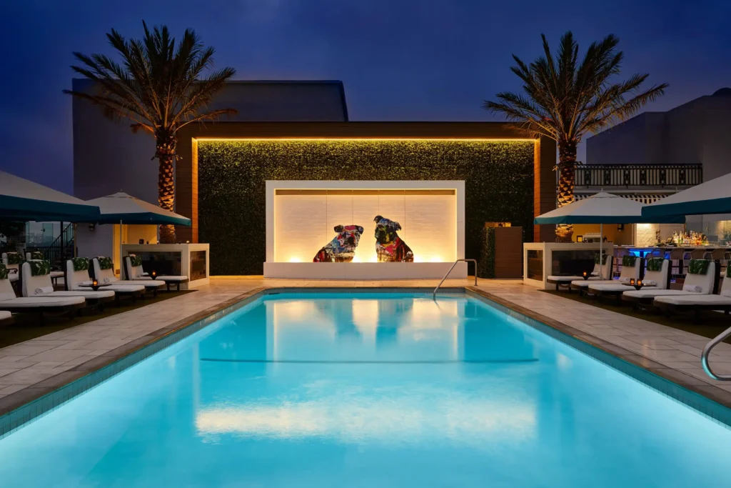 Evening rooftop pool scene with softly illuminated turquoise water, surrounded by neatly arranged lounge chairs and blue-trimmed umbrellas. Two palm trees frame a central backlit art feature set into a green wall, while warm ambient lighting and a nearby bar create a refined, upscale nighttime atmosphere