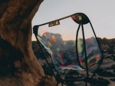 Lightweight Helinox x Gramicci camping chair with a translucent, iridescent seat, positioned on rocky terrain inside a natural stone arch, with a desert landscape and soft sunset tones visible through the material