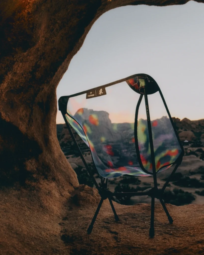 Lightweight Helinox x Gramicci camping chair with a translucent, iridescent seat, positioned on rocky terrain inside a natural stone arch, with a desert landscape and soft sunset tones visible through the material