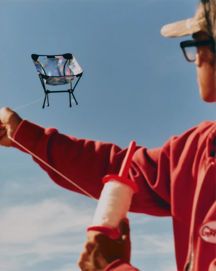 Person in a red jacket and cap holding a spool of string, flying a lightweight Helinox x Gramicci camping chair like a kite against a clear blue sky