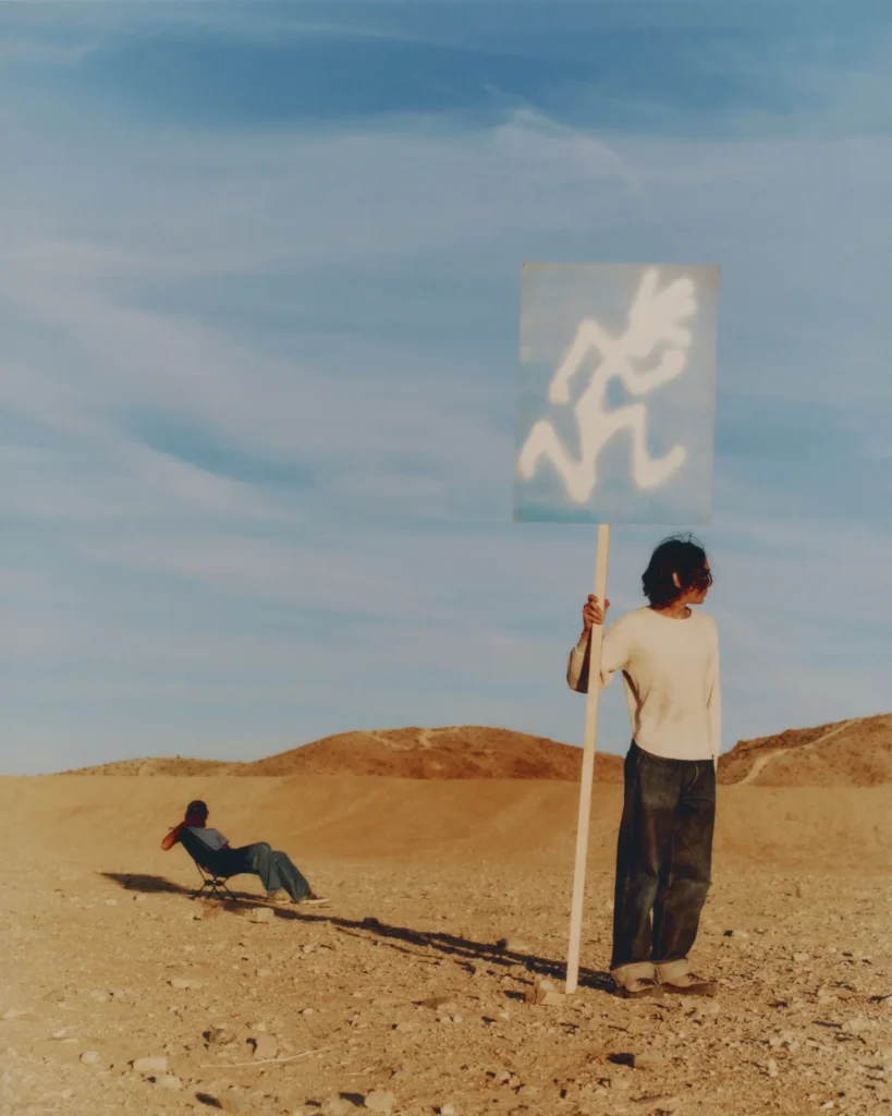 Person standing in a sunlit desert landscape holding a tall sign with the Gramicci logo, while another figure relaxes in a low camping chair in the background under a wide, cloud-streaked sky