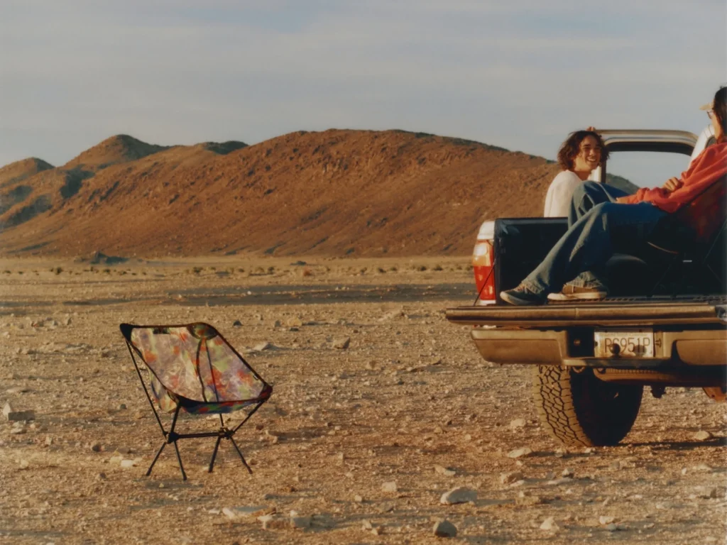 Person seated in a translucent Helinox x Gramicci camping chair strapped to the roof of a moving pickup truck, raising an arm as another passenger rides in the truck bed across a wide, arid landscape