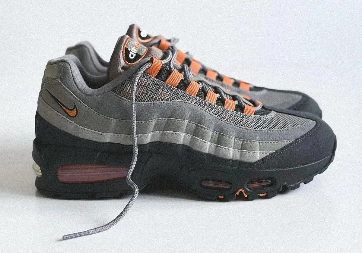A pair of Nike Air Max 95 sneakers in gradient grey layers with black mudguards and orange lace loops, shown in a clean studio setting with one lace loosely draped, highlighting the mesh upper, mini Swoosh, and visible Air cushioning units in the sole