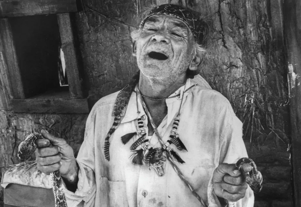 An elderly man with a headband and layered necklaces holds two snakes while standing against a textured wall in a black-and-white scene