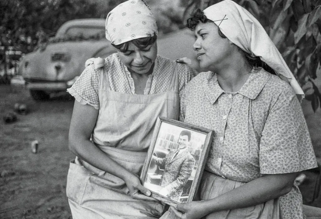Two women in headscarves stand closely together outdoors, holding a framed photograph, in an emotional black-and-white scene