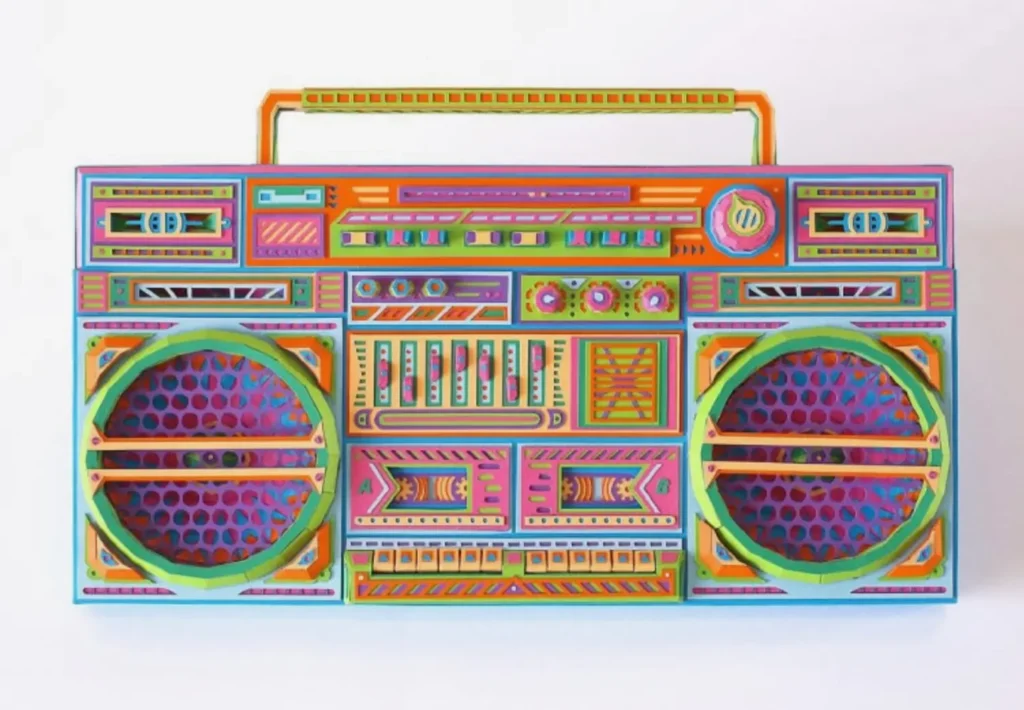 Handmade paper boombox sculpture featuring vibrant multicolor panels, layered speaker grilles, and intricate cut-and-fold details, recreating a vintage stereo system with precise geometric patterns and dimensional craftsmanship