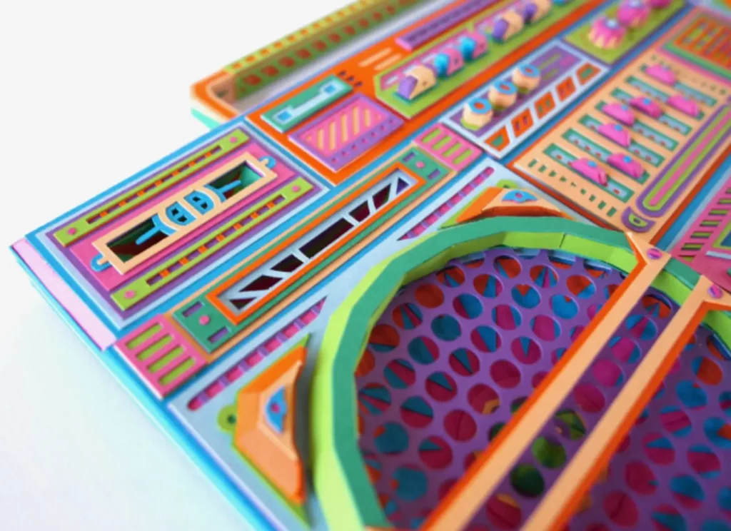 Close-up of a handmade paper boombox sculpture showing layered speaker grille, geometric control panels, and multicolored cut-and-fold details, with precise paper construction creating depth and texture across buttons, sliders, and perforated speaker patterns