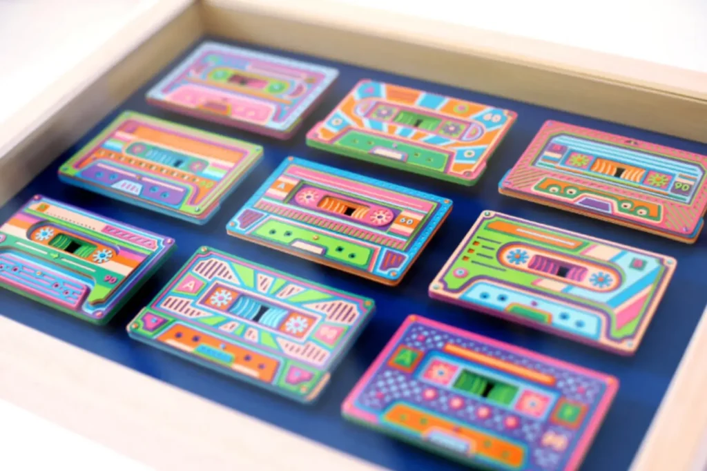 Colorful handmade paper cassette tape sculptures arranged in a grid within a shadow box frame, featuring layered geometric details, bright pastel tones, and intricate cut-and-fold craftsmanship reminiscent of vintage audio design