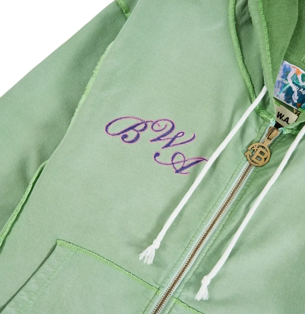 Better With Age Magic Zip-Up Hoodie close-up showing green washed cotton fleece, purple embroidered BWA logo on chest, exposed raw seams, white drawstrings, and gold zipper with custom branded pull