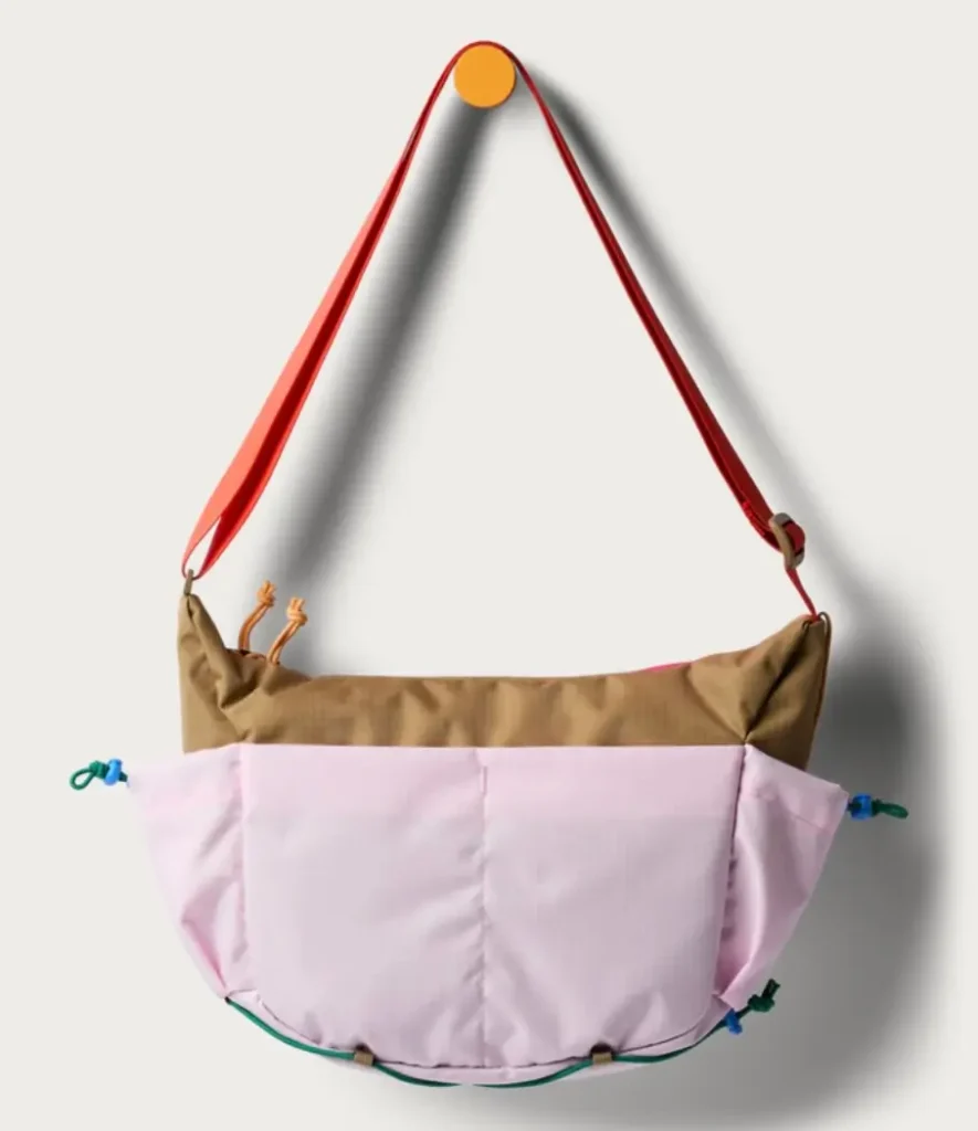 Back view of the Cotopaxi x Free People shoulder bag in ginger and powder pink, featuring a wide red adjustable strap, ripstop fabric texture, and colorful cord details along the sides and base