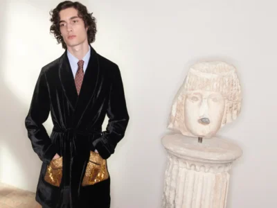 A male model with wavy dark hair stands against a minimal backdrop wearing a black velvet robe-style jacket over a shirt and patterned tie, paired with gold textured trousers, positioned beside a carved stone pedestal topped with a sculptural head