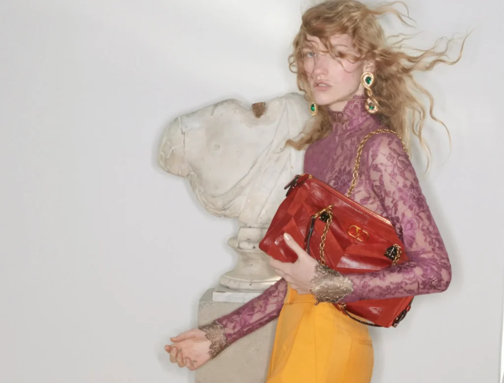 A female model with windswept blonde hair wears a sheer purple lace top and mustard-toned bottoms, holding a structured red leather handbag with gold chain detailing, posed next to a classical stone bust on a pedestal