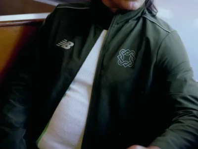 Man in dark green jacket sitting at a café table with a cup of coffee, looking off to the side