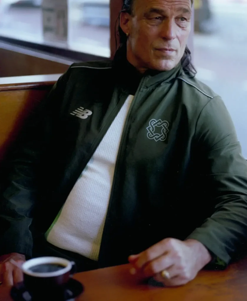 Man in dark green jacket sitting at a café table with a cup of coffee, looking off to the side