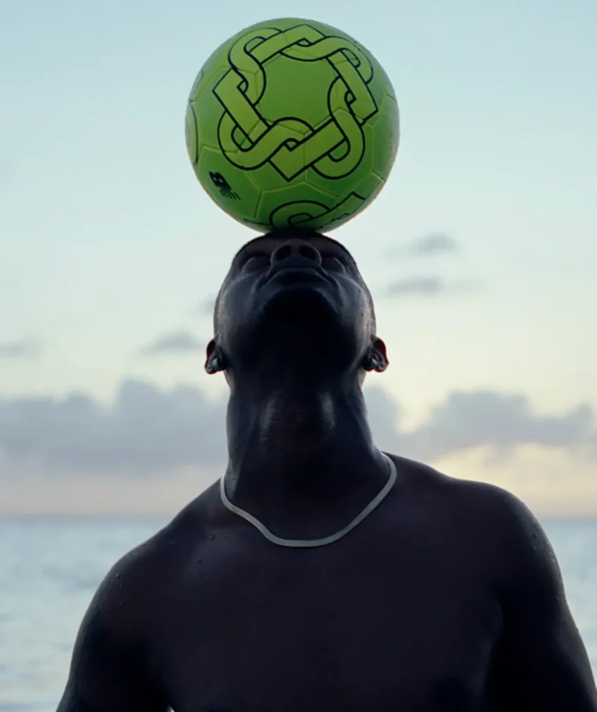 Person balancing a bright green soccer ball on their forehead by the ocean at dusk
