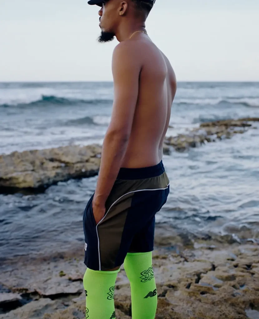Shirtless person in black shorts and neon green socks standing on a rocky shoreline by the ocean