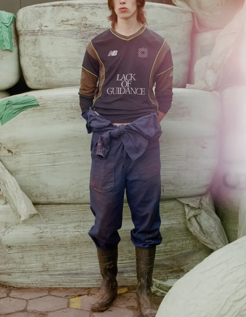 Person wearing a dark “Lack of Guidance” jersey, loose work pants, and rubber boots standing in front of stacked containers