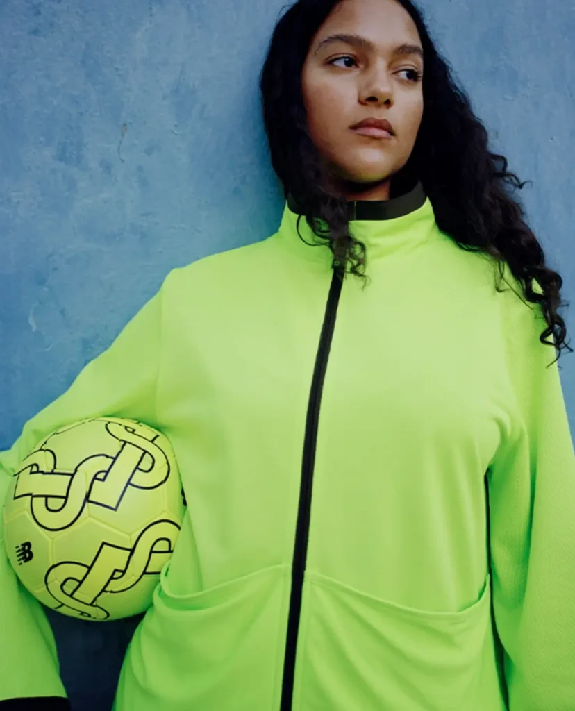 Person in a neon green jacket holding a matching soccer ball, standing against a blue wall.