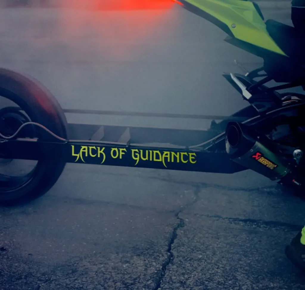 Close-up of a motorcycle rear with “Lack of Guidance” text on the frame, surrounded by tire smoke on the road