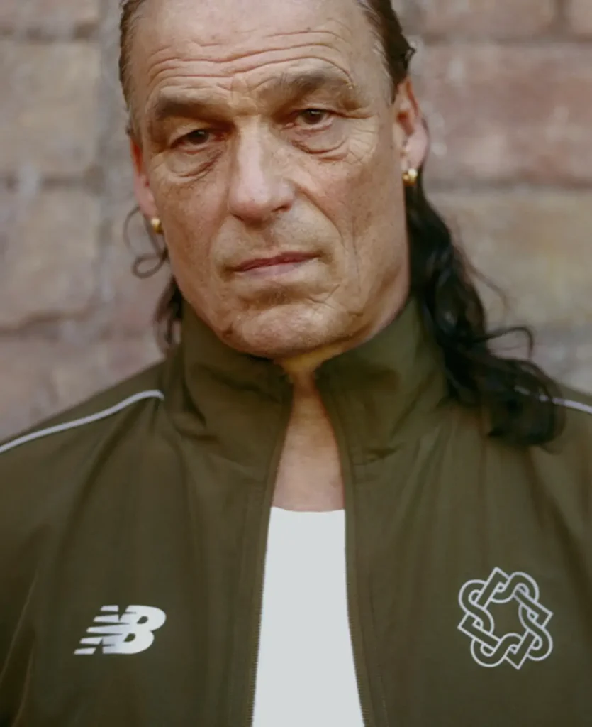 Close-up portrait of a man in a dark green New Balance jacket with a “Lack of Guidance” logo, standing against a textured wall