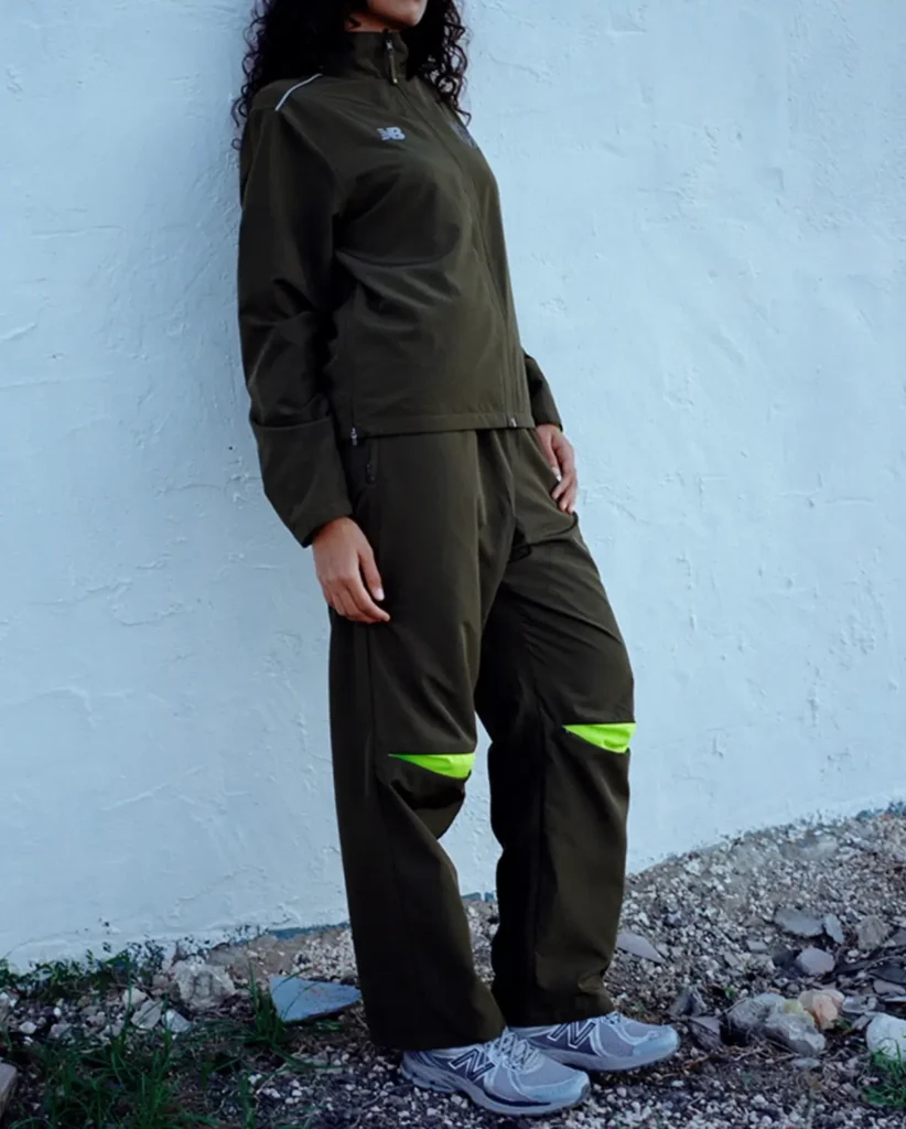 Person wearing an olive green tracksuit with neon accents and New Balance sneakers, standing against a light wall
