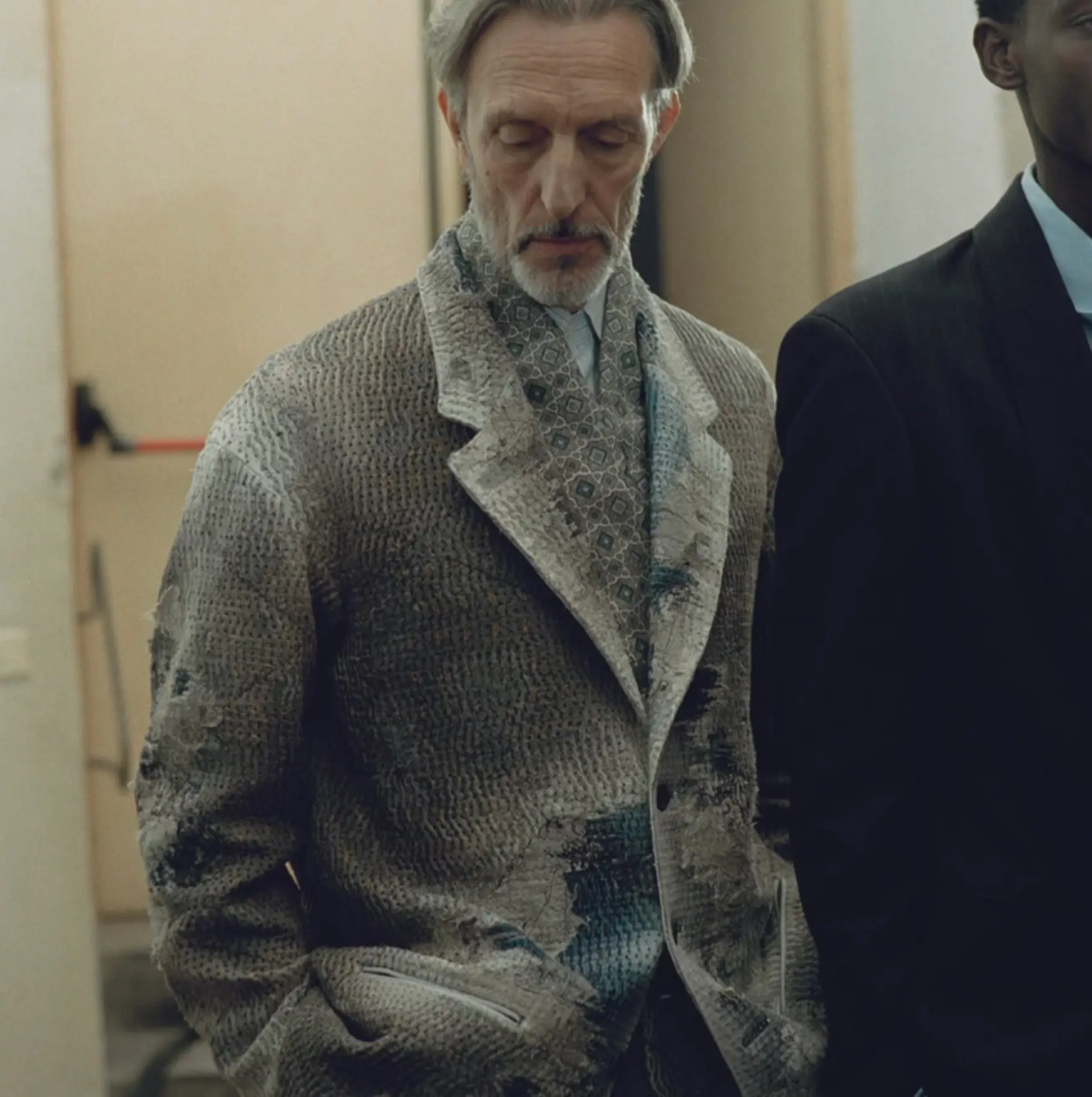 An older man with graying hair and a short beard looks downward in a softly lit interior, dressed in a textured, patterned overcoat layered over a coordinating shirt and tie. The muted tones and intricate fabric details give the outfit a refined, artisanal feel, while his relaxed posture and hands-in-pocket stance convey quiet sophistication