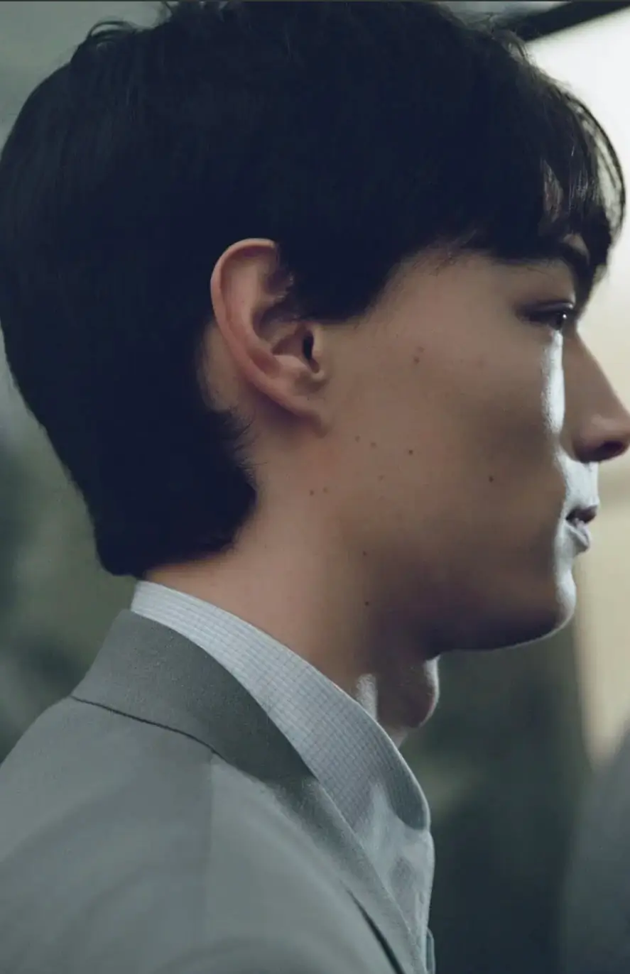 A close-up side profile of a young man with short, dark hair, wearing a light-toned suit jacket over a subtly textured shirt. The soft, natural lighting highlights his facial contours and calm, introspective expression, giving the image a quiet, refined mood