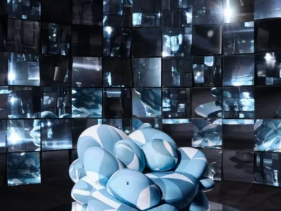 Futuristic installation featuring a sculptural, cloud-like blue seat set on a reflective platform, surrounded by a wall of mirrored panels that fragment light and reflections in a dark, immersive space