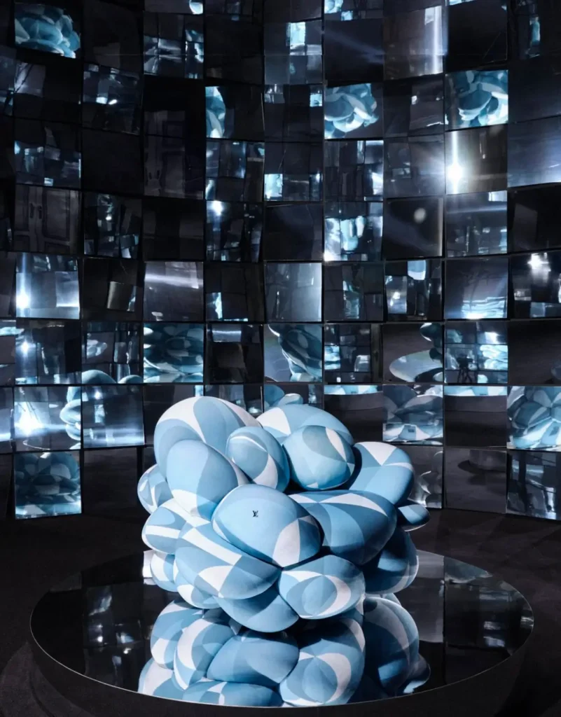 Futuristic installation featuring a sculptural, cloud-like blue seat set on a reflective platform, surrounded by a wall of mirrored panels that fragment light and reflections in a dark, immersive space