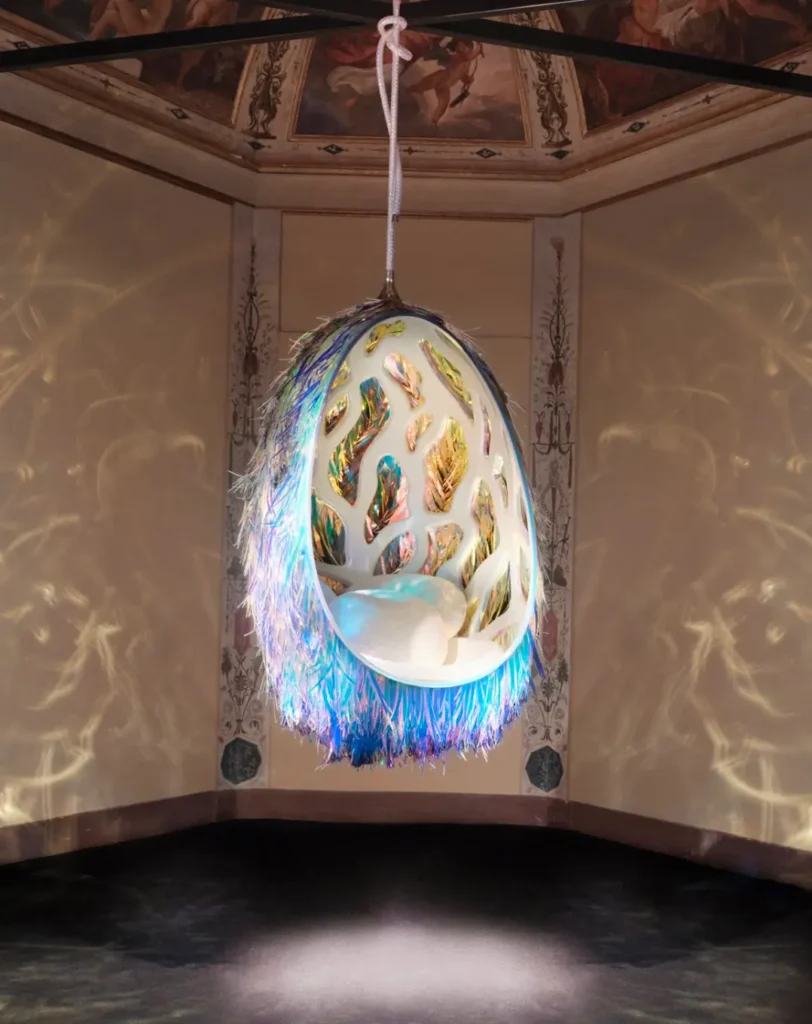 Suspended cocoon-like sculptural seat with iridescent interior panels and fringe detailing, glowing with prismatic light inside an ornate frescoed room, casting reflective patterns across the walls