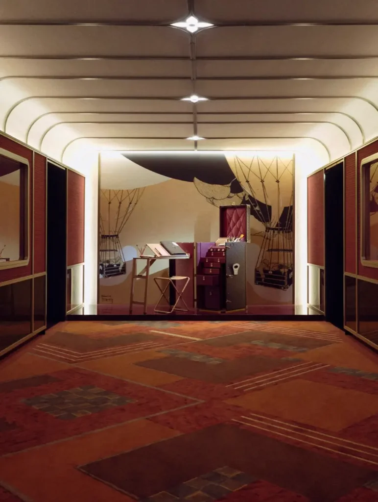Symmetrical gallery corridor with warm wood paneling and soft overhead lighting, leading to a central display of luxury trunks and travel accessories set against a graphic mural backdrop