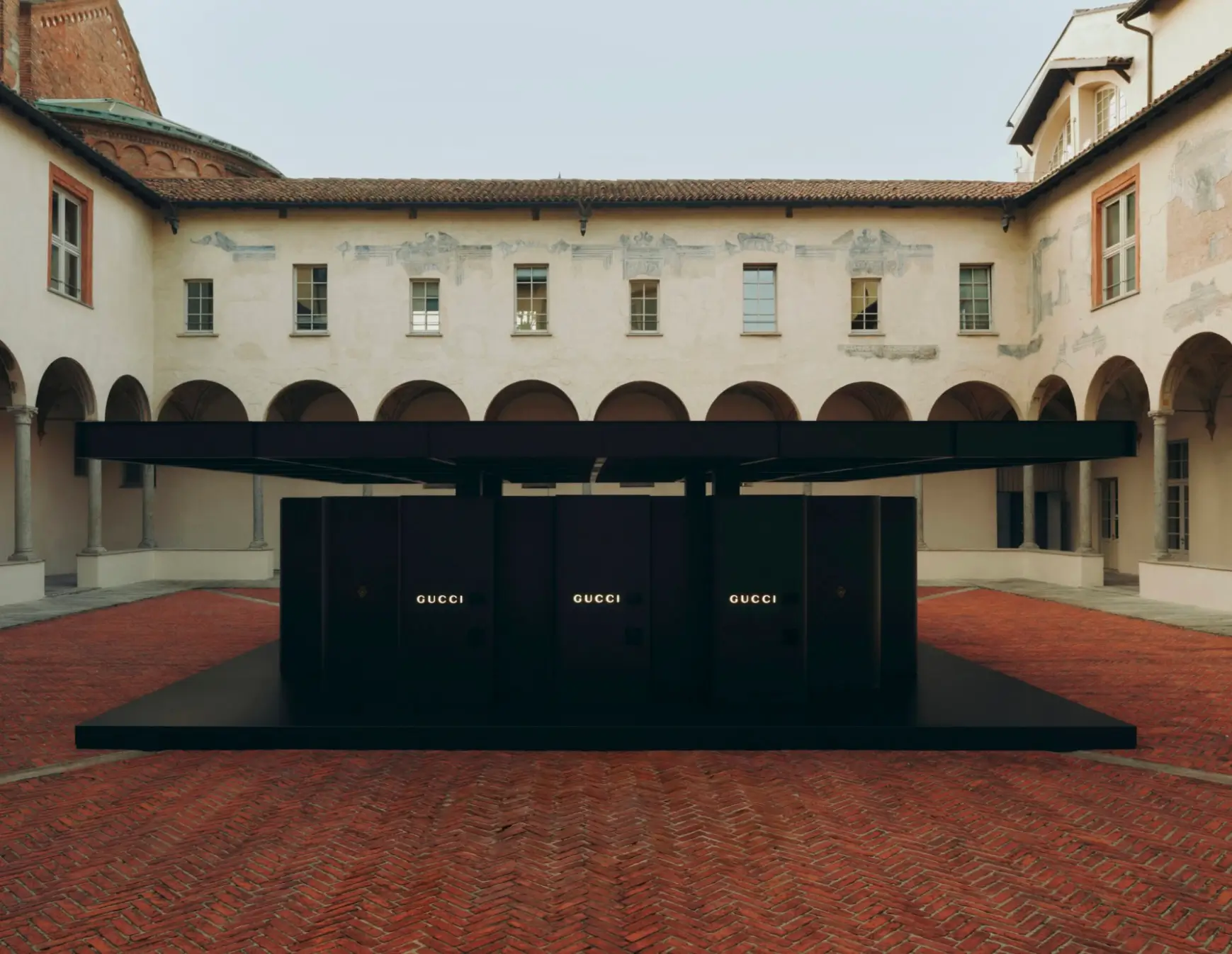 Minimalist Gucci installation featuring a large black canopy structure and branded panels, set within a historic courtyard with arched colonnades and brick flooring