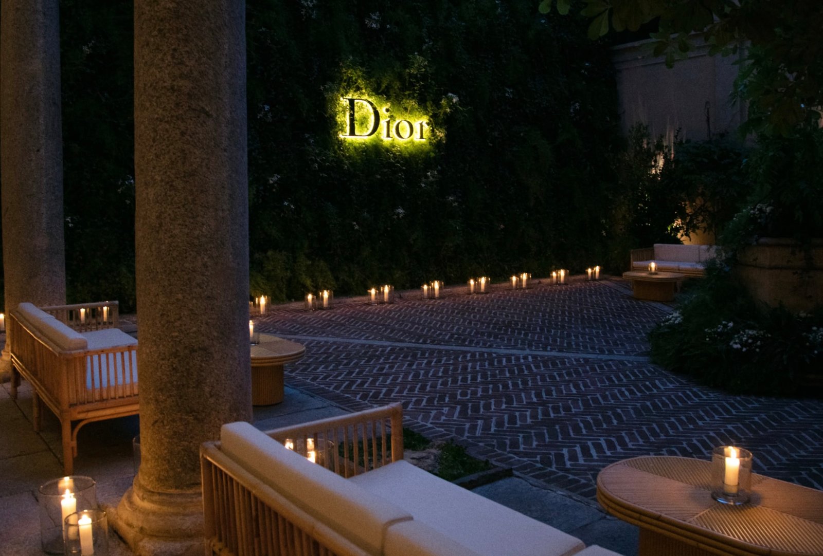 Evening courtyard setting with a glowing Dior logo set against lush greenery, illuminated by rows of candles and surrounded by relaxed lounge seating on a brick-paved terrace