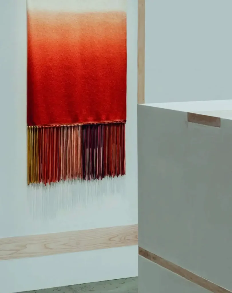 A minimalist gallery corner showcasing a textile artwork with a rich gradient transitioning from soft beige to deep, saturated red. The piece hangs vertically against a clean white wall, its lower edge finished with long, delicate fringe strands in varying tones of red, rust, and amber, adding movement and tactile depth. Adjacent to it, a sculptural white display plinth with subtle wood inlays reinforces the exhibition’s refined, material-focused aesthetic. The composition is quiet and intentional, emphasizing craftsmanship, texture, and the interplay between color, fabric, and space
