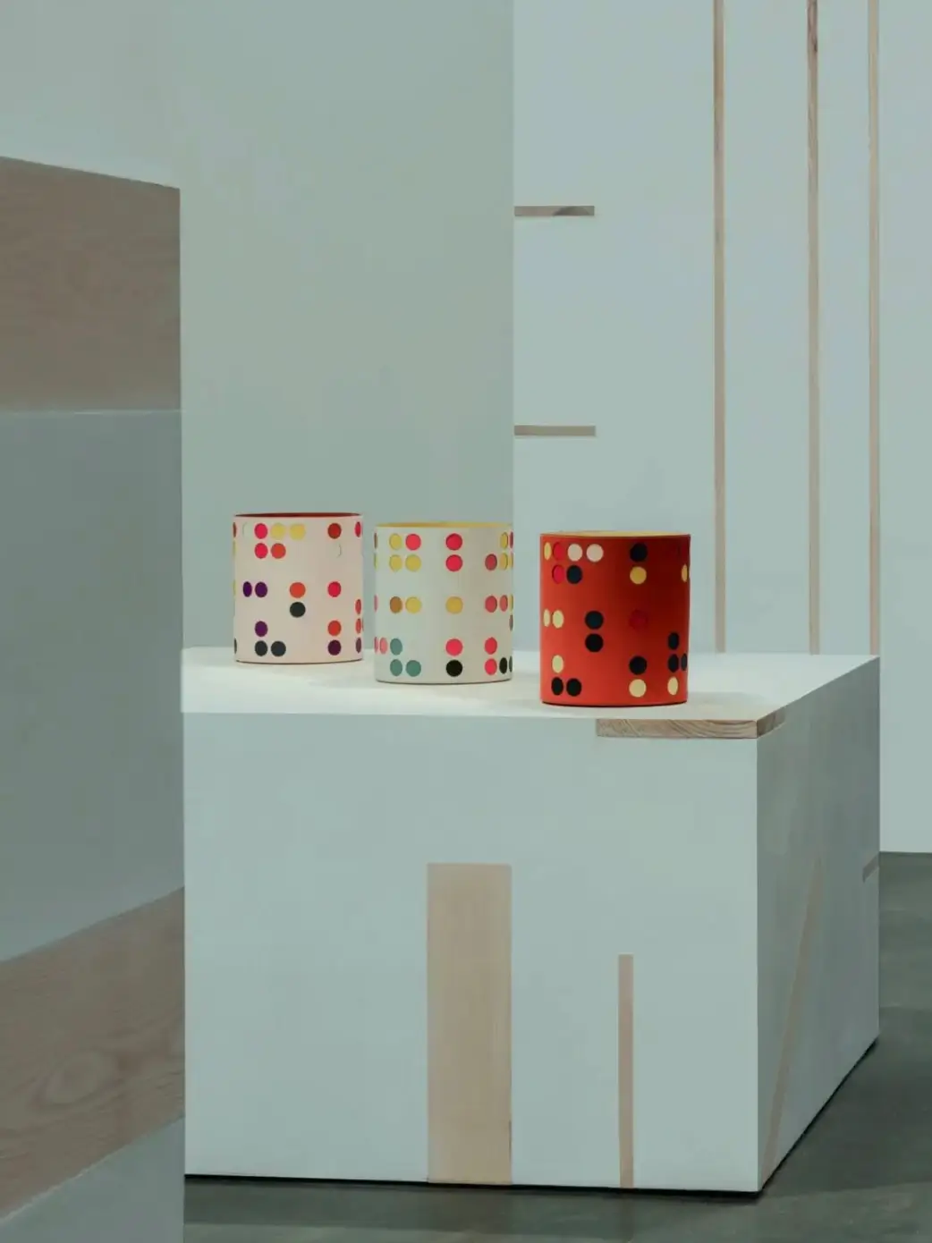 Three cylindrical objects with colorful polka-dot patterns displayed on a white pedestal within a minimalist gallery space featuring clean lines and light wood detailing