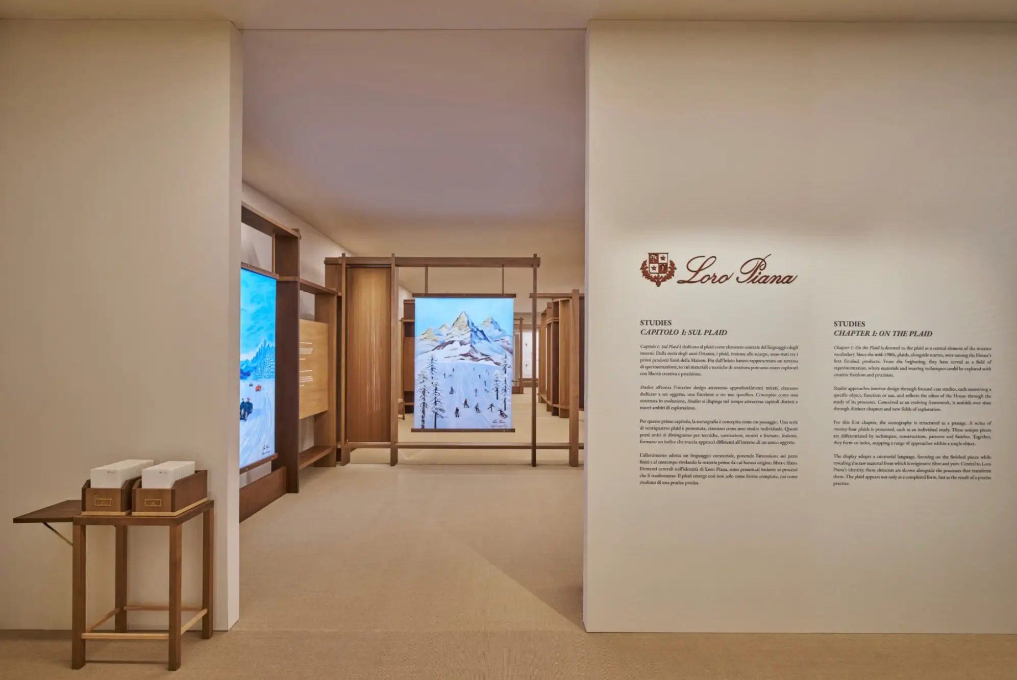 A refined exhibition entrance space featuring warm, minimalist architecture with soft beige walls and light carpeting. On the right, a clean wall display presents the Loro Piana crest and logo above neatly typeset curatorial text, introducing the exhibit. Beyond the opening, a sequence of wooden display frames creates a gallery corridor, showcasing textile-inspired artworks, including a central panel depicting a snowy mountain landscape. To the left, a small wooden table holds neatly arranged boxed items, reinforcing the brand’s emphasis on craftsmanship and material heritage. The overall atmosphere is quiet, curated, and immersive, blending luxury retail with museum-like presentation