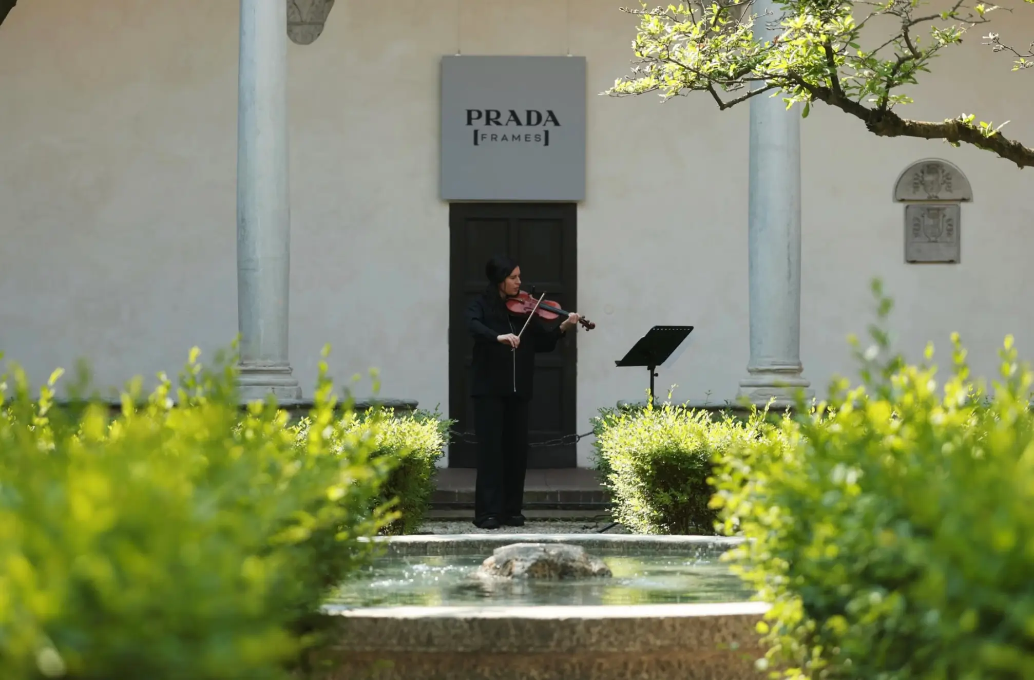 A serene courtyard performance framed by classical architecture and greenery, centered on a violinist dressed in black, mid-performance before a discreet audience setup. Above the doorway behind them, a sign reads Prada Frames, indicating a cultural or intellectual program rather than a conventional fashion presentation. The composition is cinematic: trimmed hedges guide the eye toward a small reflective fountain in the foreground, while stone columns and a pale façade create a quiet, almost monastic backdrop. The musician stands alone, focused, with a music stand nearby—suggesting an intimate recital rather than spectacle. The atmosphere feels intentional and restrained—luxury expressed through silence, space, and cultural alignment. It’s less about product, more about positioning: art, sound, and environment woven into the identity of Prada as a curator of thought as much as design